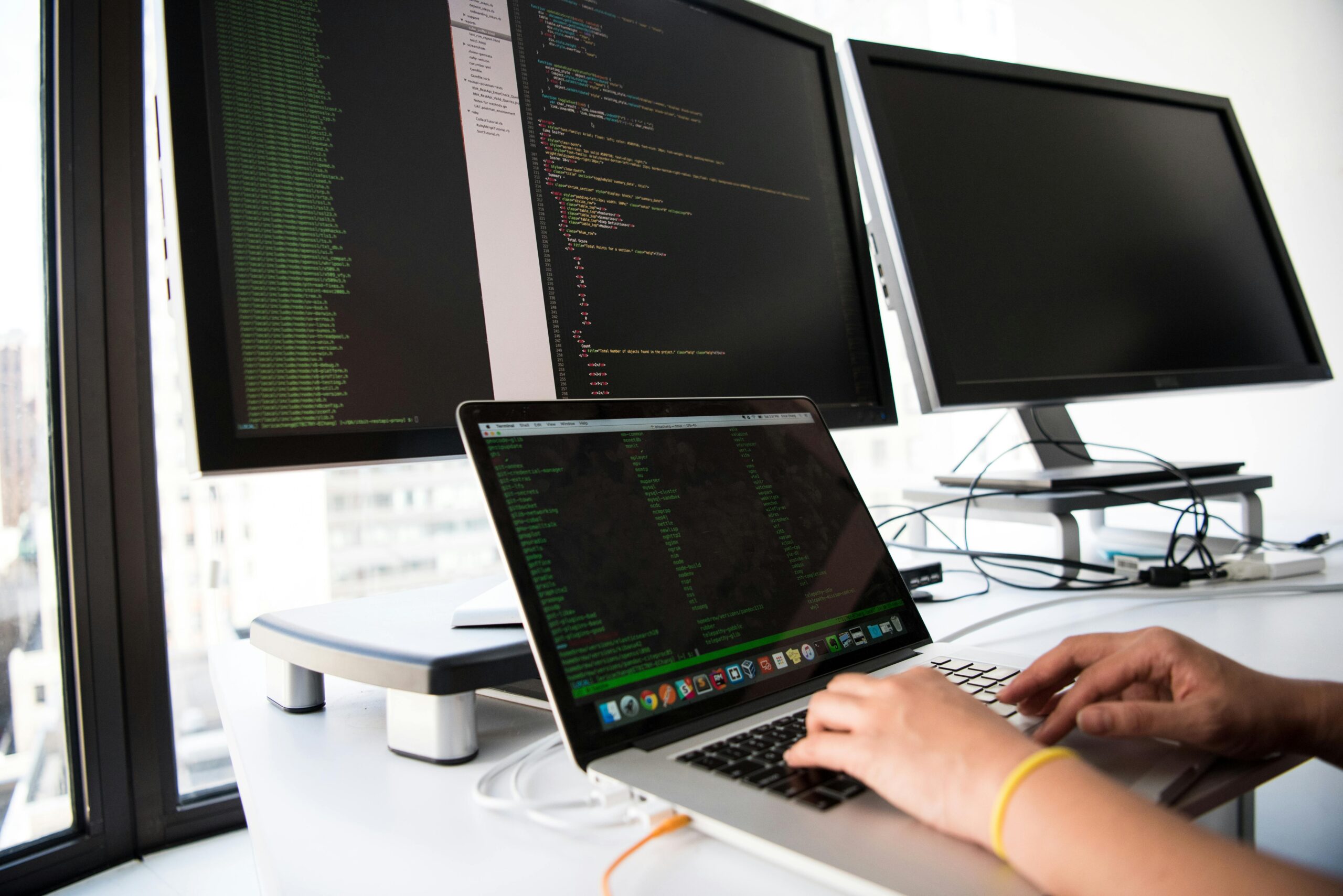 Home A developer writes code on a laptop in front of multiple monitors in an office setting.